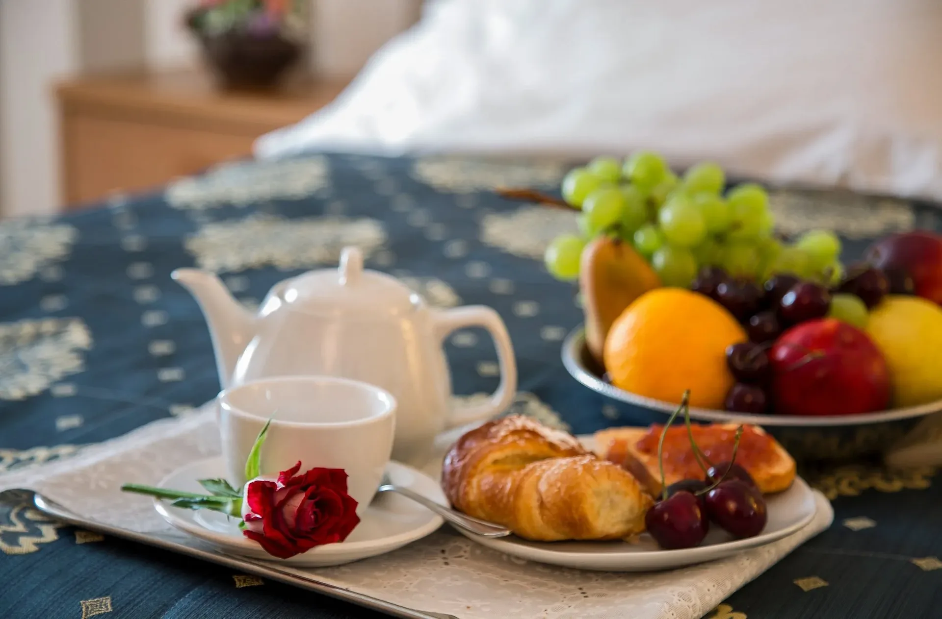 Vassoio colazione con teiera, cornetto, frutta e rosa rossa sul letto