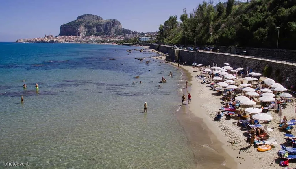 Spiaggia sabbiosa con ombrelloni e mare cristallino, con la rocca di Cefalù sullo sfondo