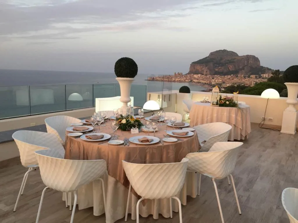 Tavolo rotondo elegantemente allestito su terrazza con vista sul mare e la rocca di Cefalù