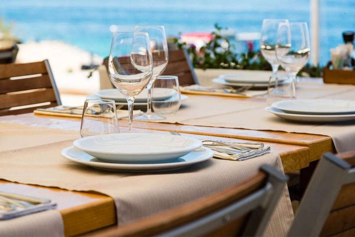 Tavolo apparecchiato di un ristorante con calici e piatti bianchi affacciato sul mare