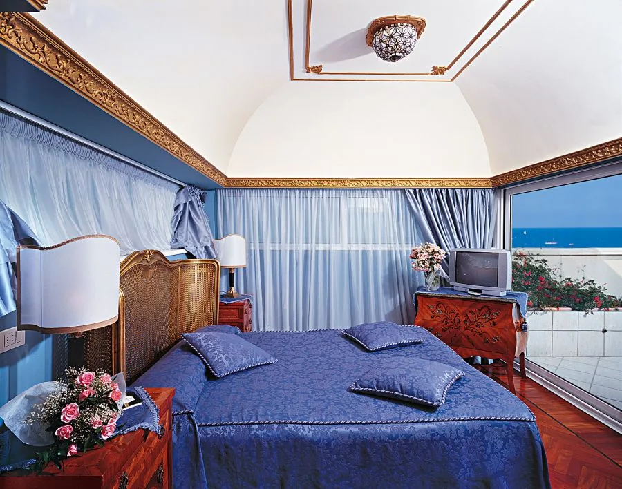 Camera con letto matrimoniale blu, soffitto a volta con cornice dorata e vista mare dalla vetrata