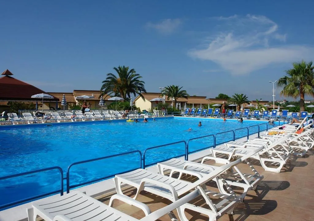 Piscina rettangolare con lettini bianchi, palme e cielo azzurro