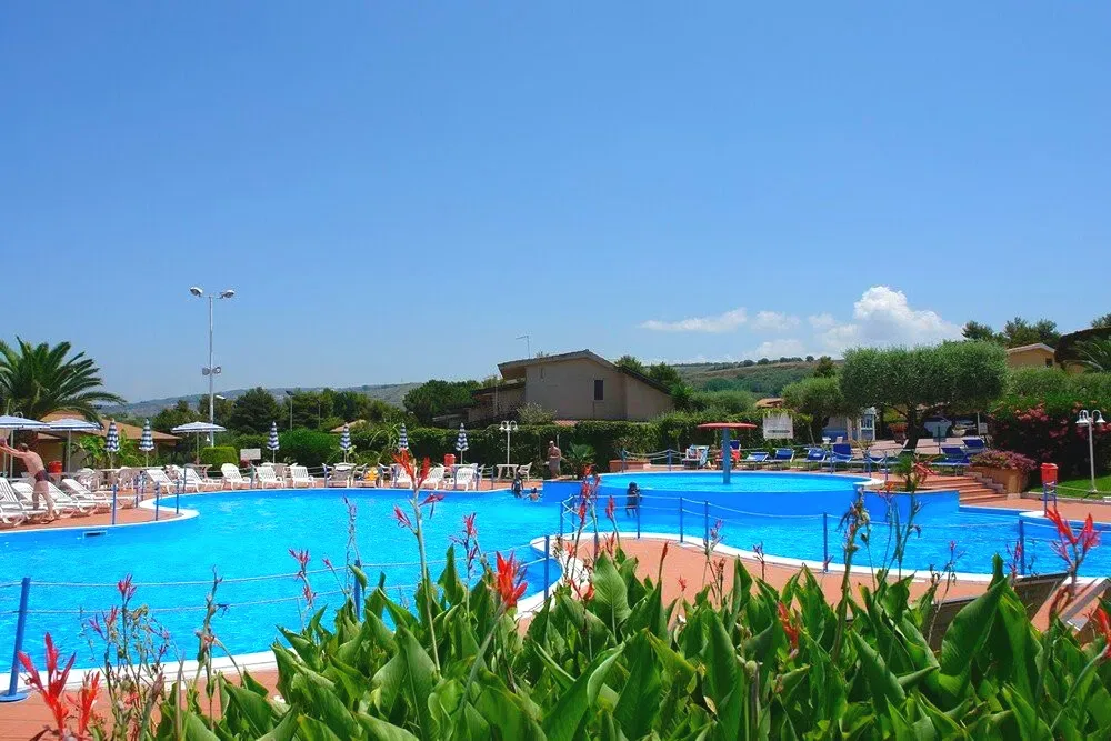 Ampia piscina esterna circondata da vegetazione e villette sotto un cielo terso