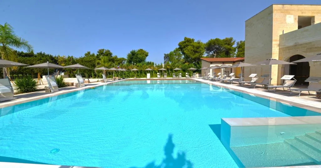 Grande piscina azzurra con scalini, ombrelloni e vegetazione mediterranea