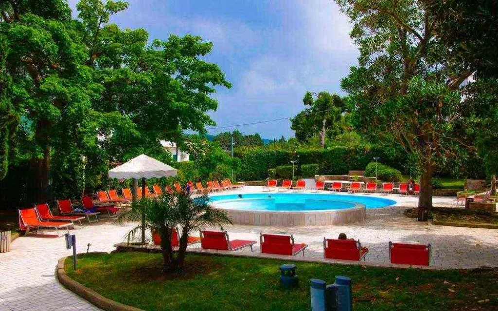 Piscina circondata da giardino con lettini rossi, ombrellone e alberi ombrosi