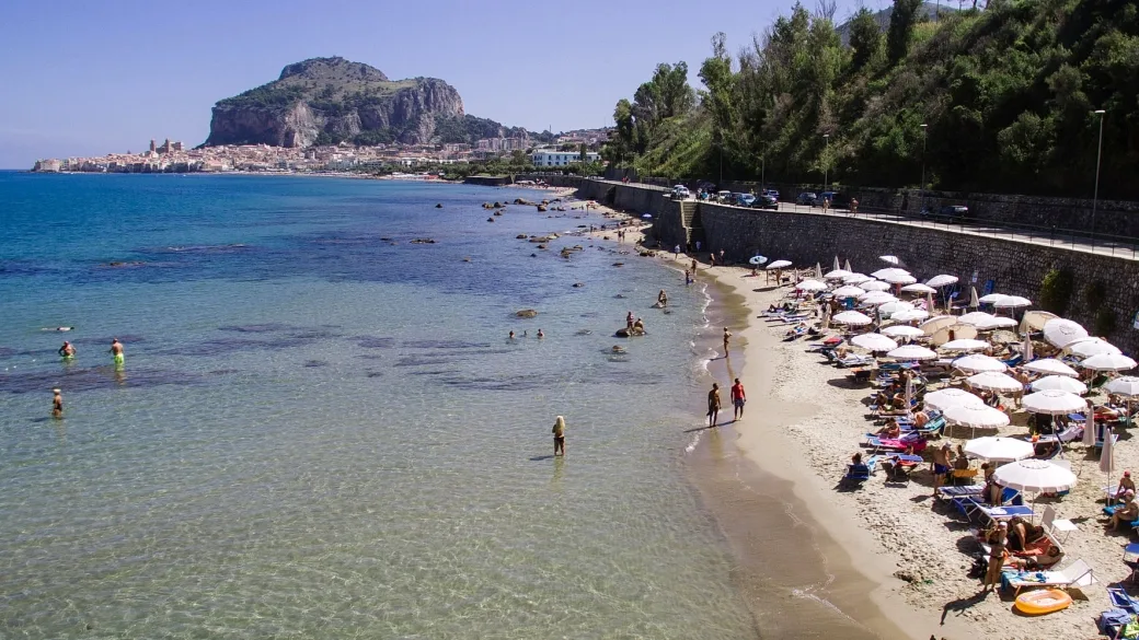 Spiaggia di sabbia con ombrelloni e mare cristallino con vista sulla rocca di Cefalù