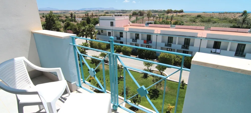 Balcone con sedie bianche in plastica e ringhiera azzurra affacciato sul giardino e gli edifici del resort