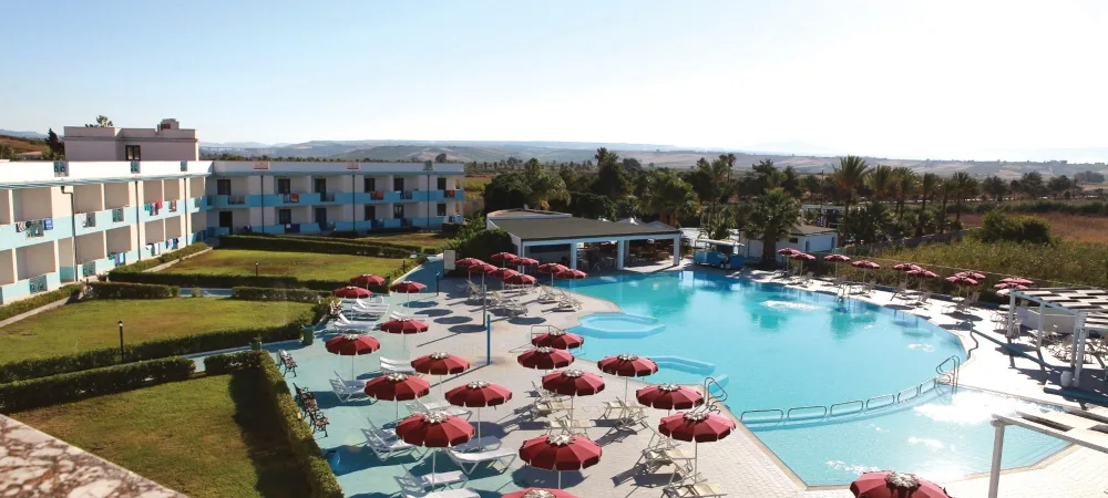 Veduta panoramica della piscina con ombrelloni rossi e lettini, edifici dell'hotel e campagna sullo sfondo