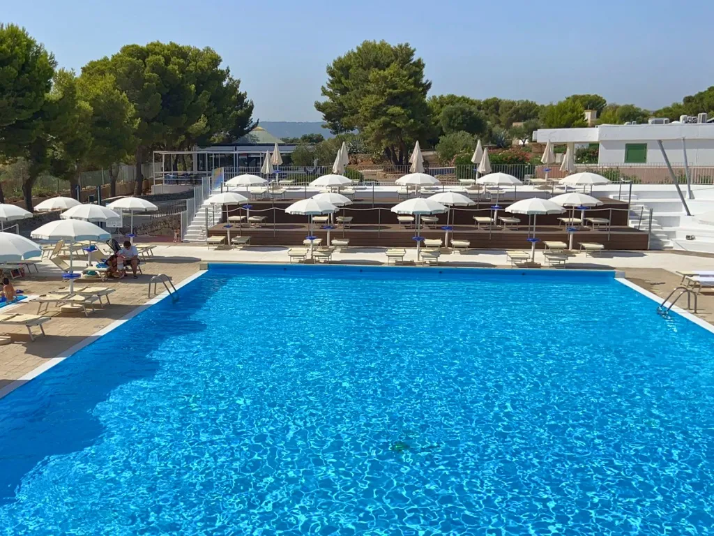 Piscina rettangolare con acqua limpida, lettini e ombrelloni bianchi circondata da pini