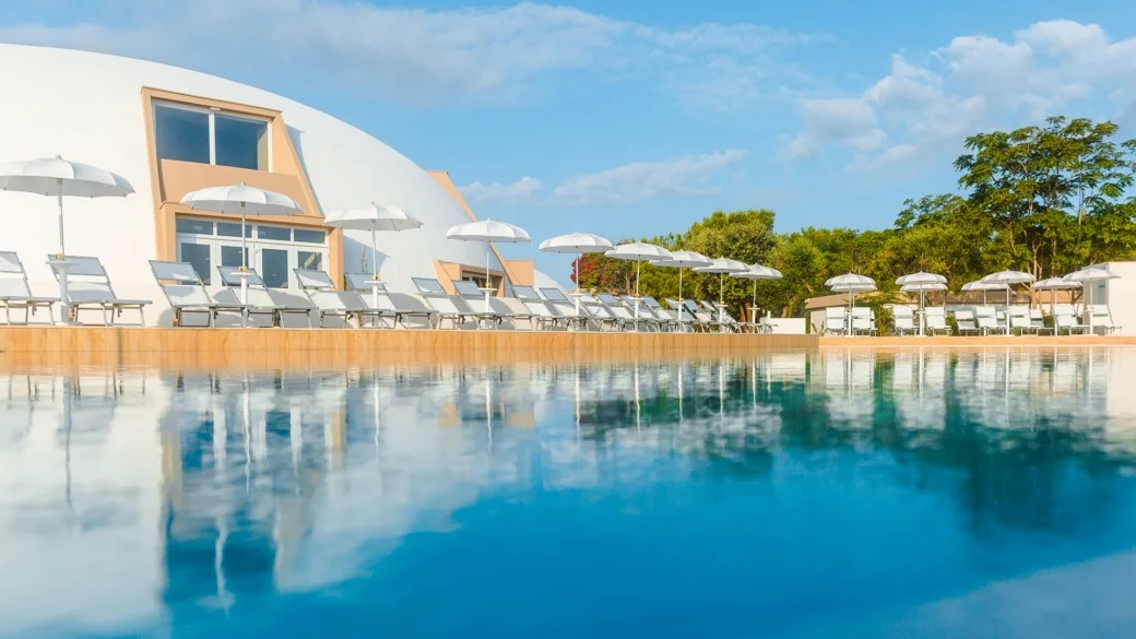 Piscina con lettini e ombrelloni bianchi accanto a edificio a cupola del resort