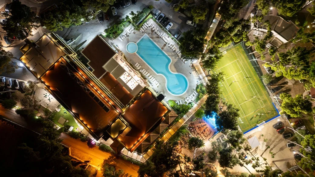 Veduta aerea notturna dell'hotel con piscina illuminata e campo sportivo