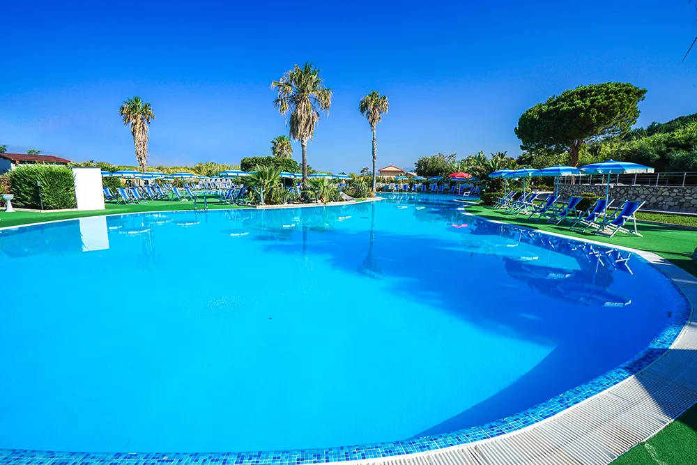 Piscina curva con acqua azzurra, lettini blu e palme sotto cielo limpido