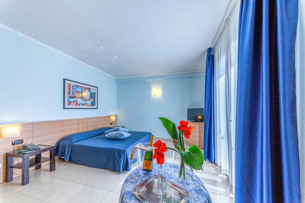 Camera matrimoniale con letto blu, ampia finestra con tende blu e tavolino con fiori e spumante