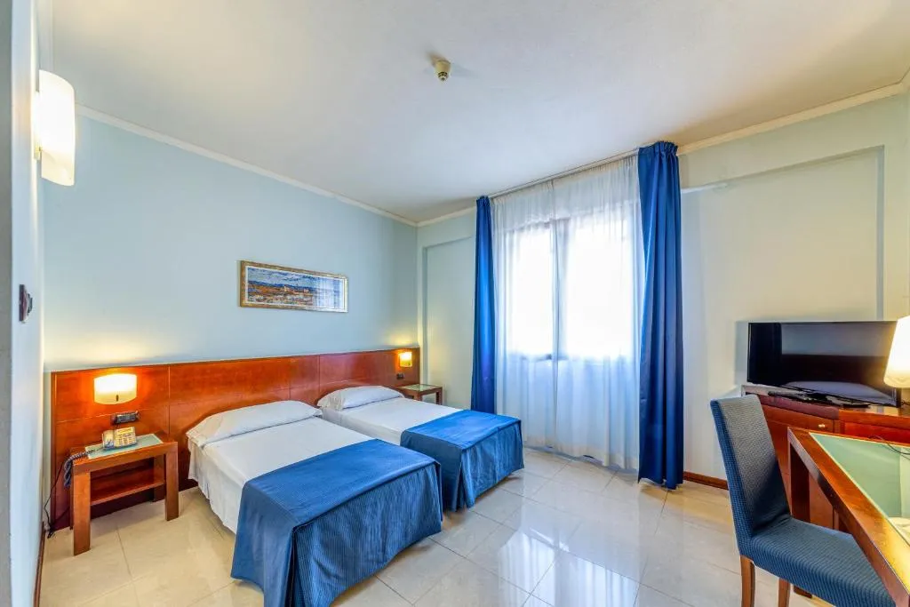 Camera doppia con due letti singoli, copriletto blu, scrivania, TV e finestra con tende blu