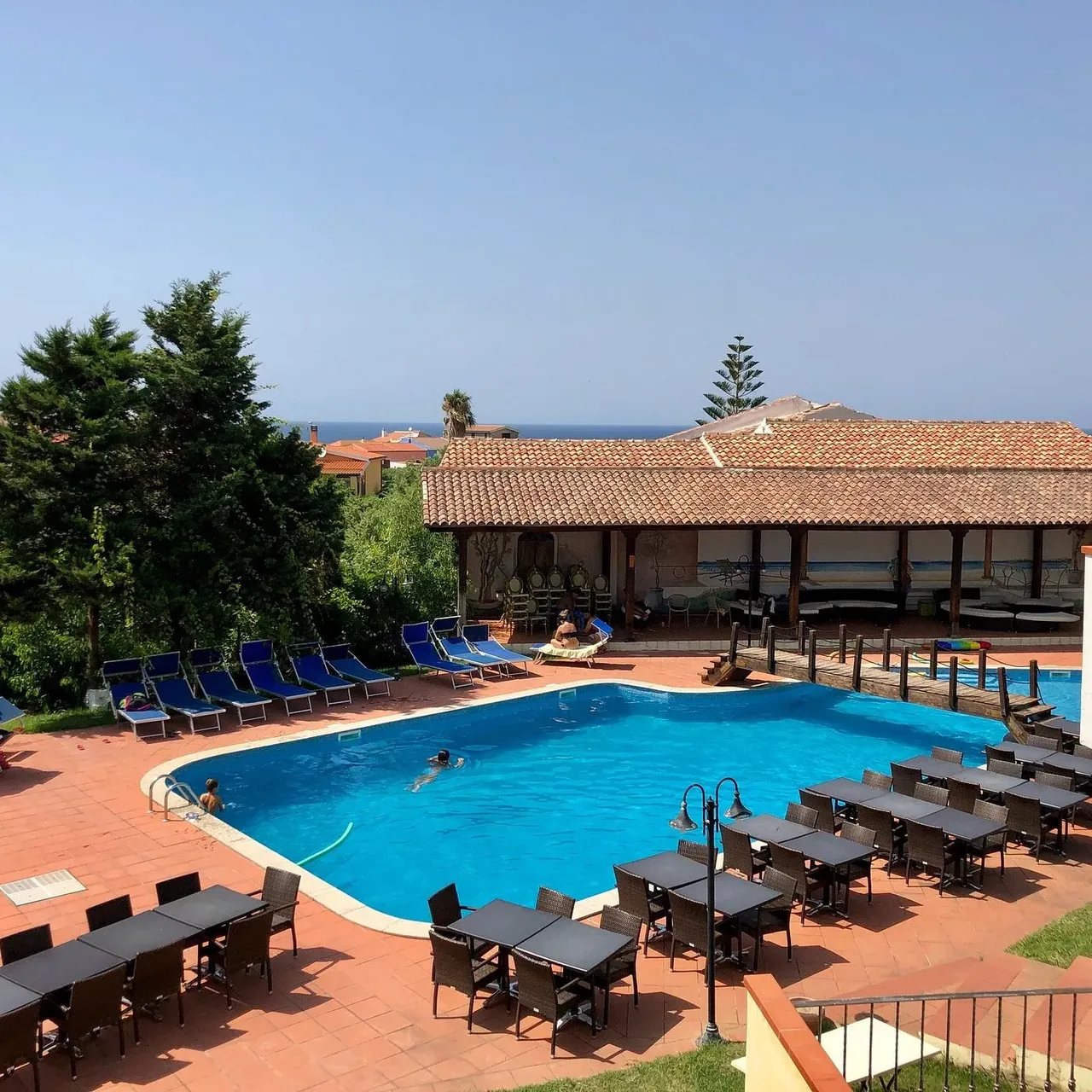 Piscina con lettini blu e tavoli del ristorante all'aperto, porticato in legno e vista mare