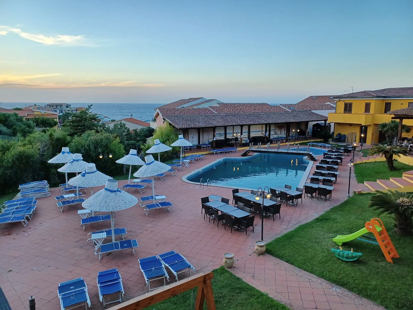 Area piscina al tramonto con ombrelloni in paglia, tavoli, area giochi per bambini e edificio giallo dell'hotel