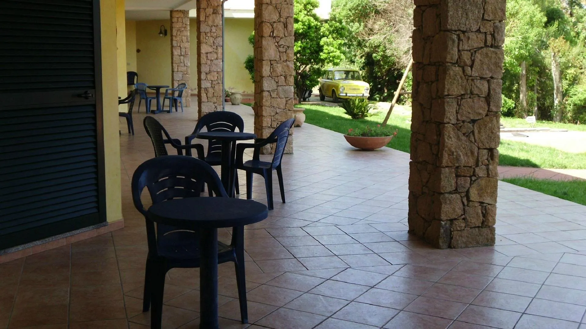 Porticato esterno dell'hotel con tavoli e sedie in plastica blu, colonne in pietra e auto d'epoca gialla sullo sfondo
