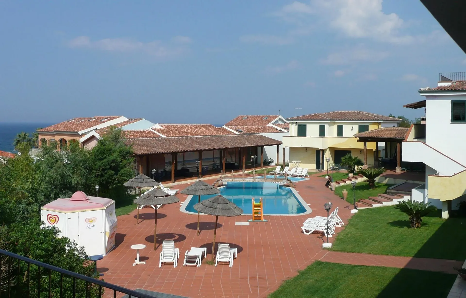 Vista dall'alto della piscina con ombrelloni in paglia, lettini e edifici dell'hotel con mare sullo sfondo