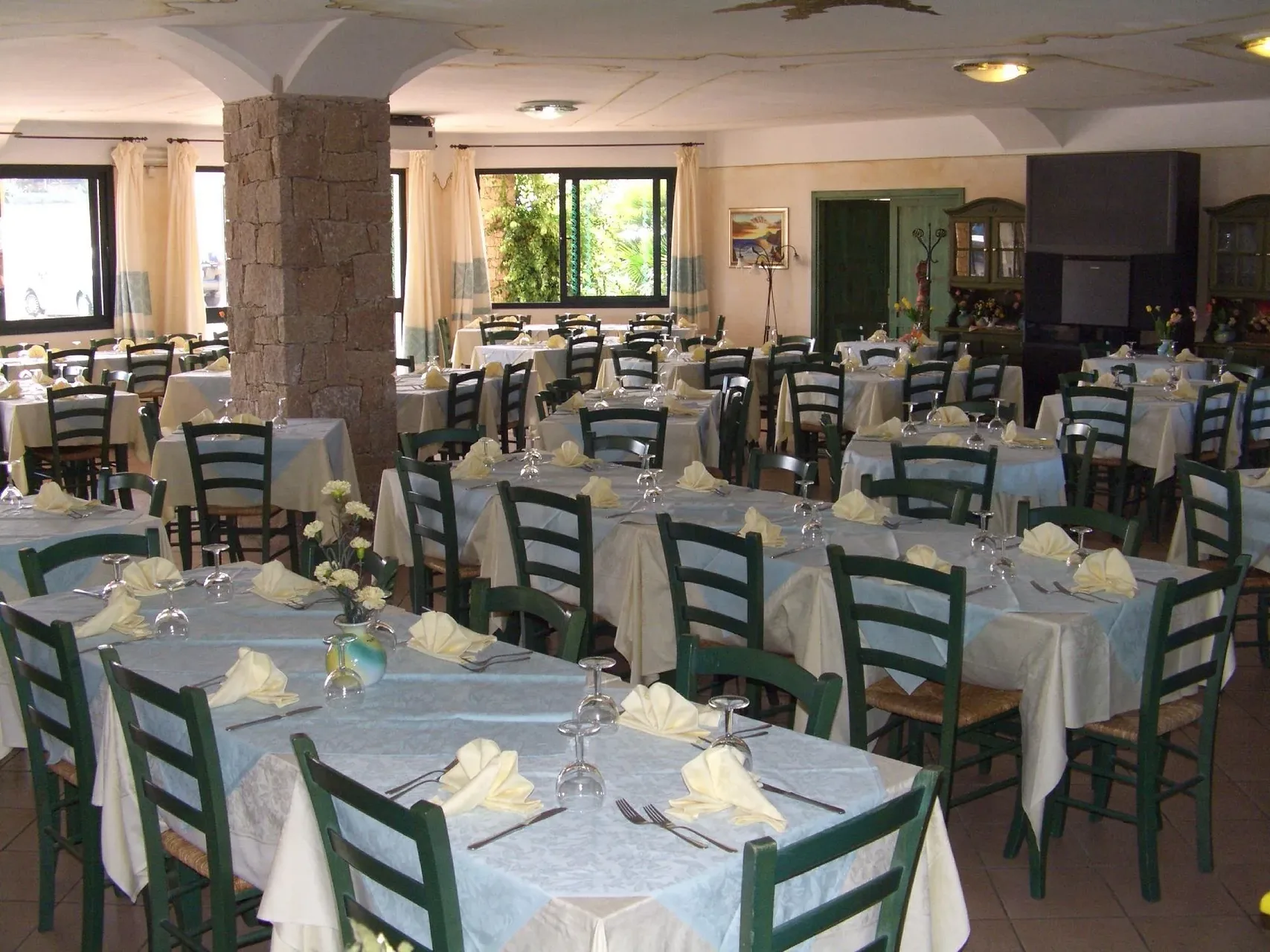 Sala ristorante interna con tavoli apparecchiati con tovaglie azzurre, sedie verdi e colonna centrale in pietra