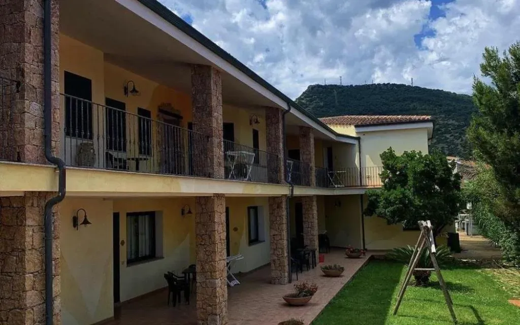 Esterno dell'hotel giallo con porticato, colonne in pietra, balconi e giardino con montagna sullo sfondo