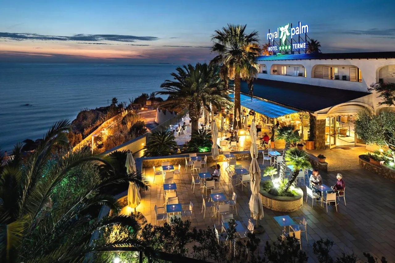 Terrazza serale del Royal Palm Hotel Terme illuminata con tavoli e palme affacciata sul mare al tramonto