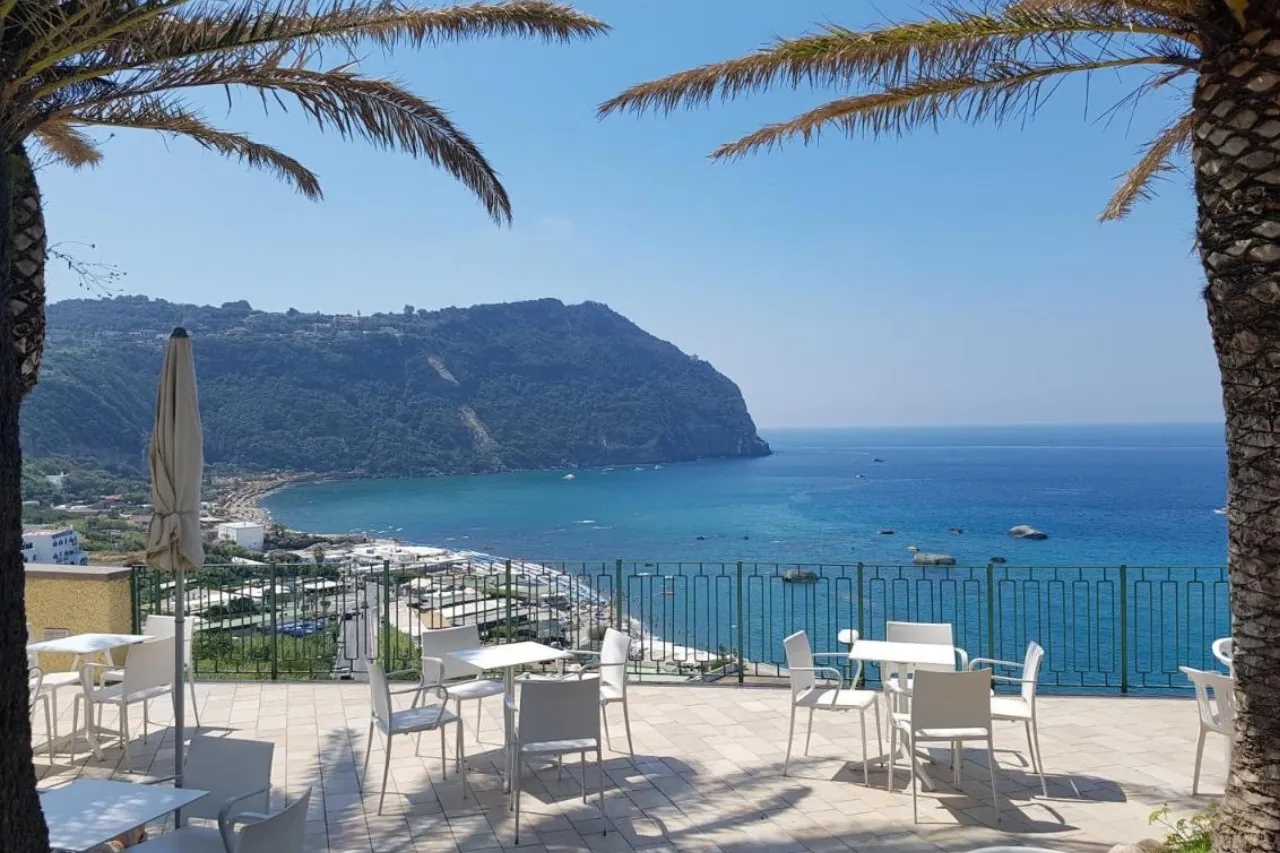 Vista panoramica sul mare e sul promontorio da una terrazza con tavoli bianchi tra le palme