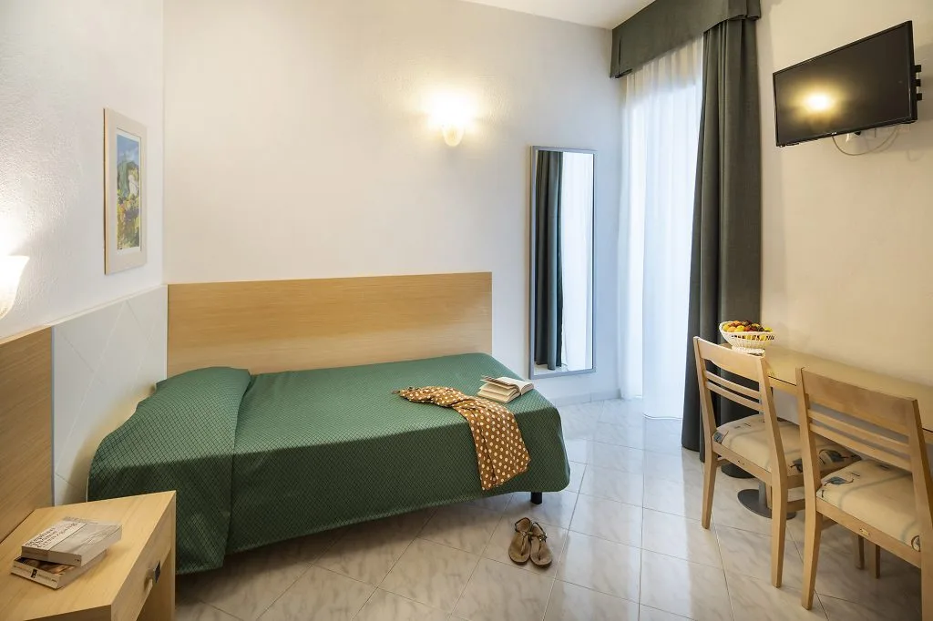 Camera singola con letto, copriletto verde, scrivania, sedie e TV a parete
