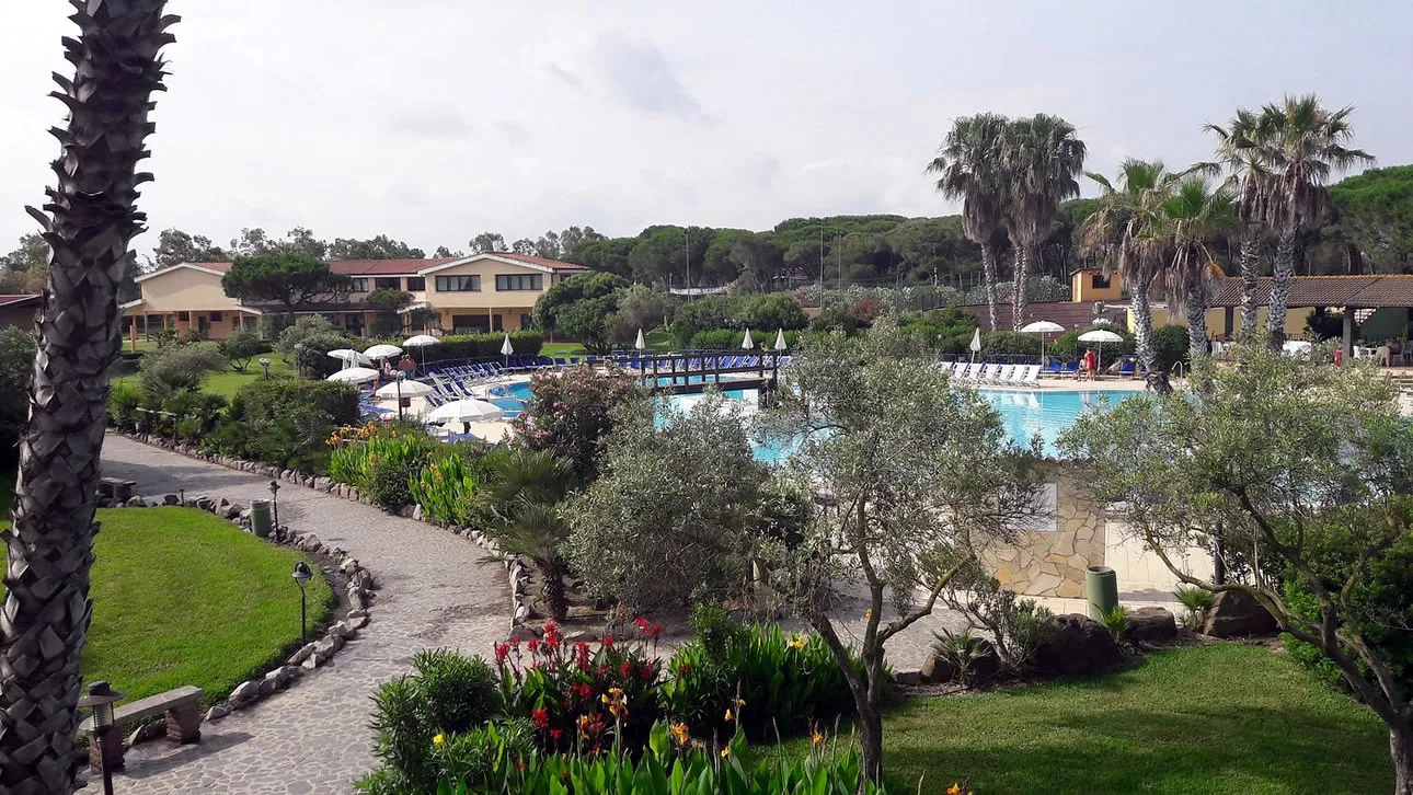 Vista sulla piscina del resort tra palme, ulivi e giardino fiorito