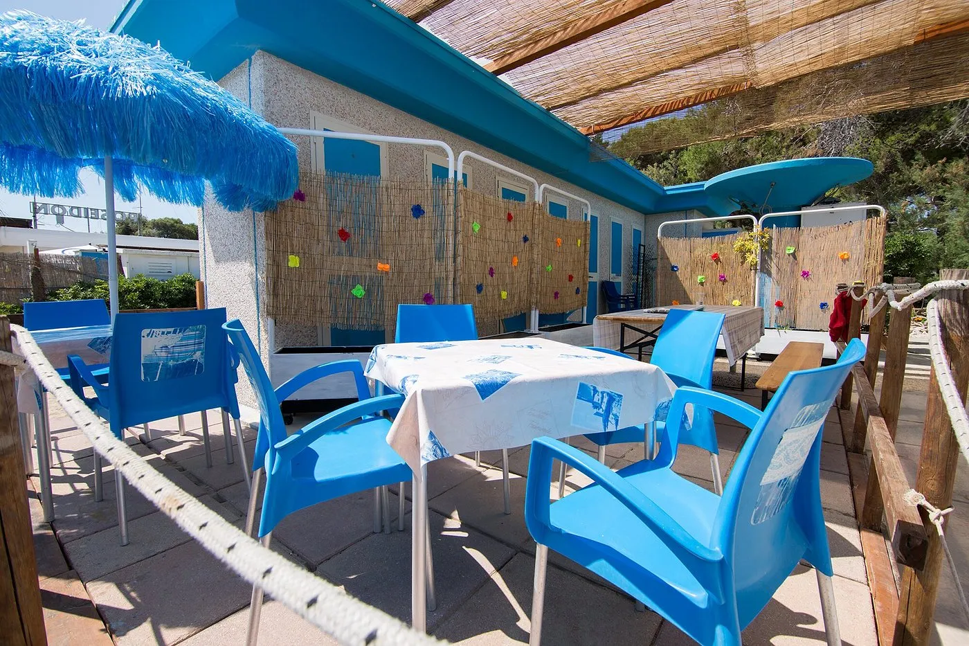 Terrazza esterna del chiosco spiaggia con tavoli, sedie azzurre e ombrellone a frange blu