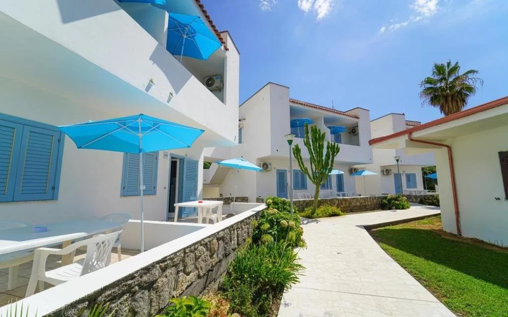 Vialetto tra gli alloggi bianchi con terrazzi, ombrelloni azzurri e giardino curato